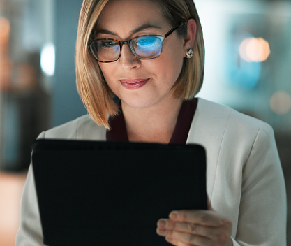 Kvinne med briller og lys jakke ser konsentrert på et nettbrett i et moderne kontormiljø. Refleksjonen i brillene antyder digitalt innhold på skjermen. Bildet illustrerer bruk av intranett for å støtte digital employee experience (DEX).