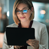 Kvinne med briller og lys jakke ser konsentrert på et nettbrett i et moderne kontormiljø. Refleksjonen i brillene antyder digitalt innhold på skjermen. Bildet illustrerer bruk av intranett for å støtte digital employee experience (DEX).